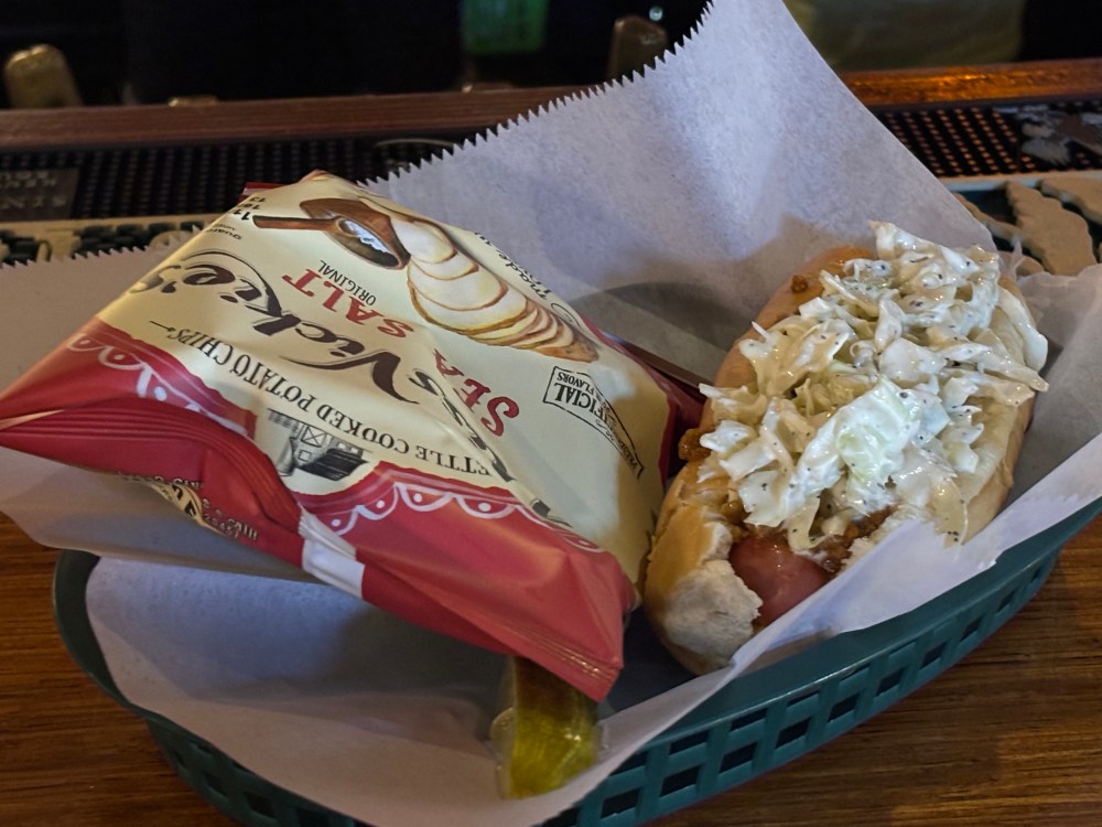 a plastic bar serving basket, with a bag of potato chips on the left and a chili slaw hot dog on the right